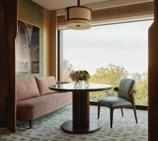 Modern dining nook with pink sofa, round glass table, and large window framing leafy treetop views 