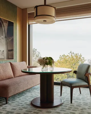 Modern dining nook with pink sofa, round glass table, and large window framing leafy treetop views 