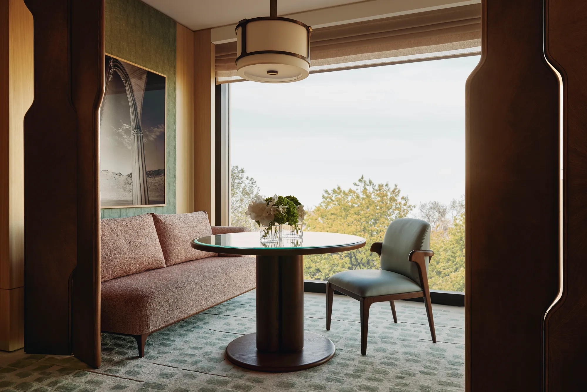 Modern dining nook with pink sofa, round glass table, and large window framing leafy treetop views 