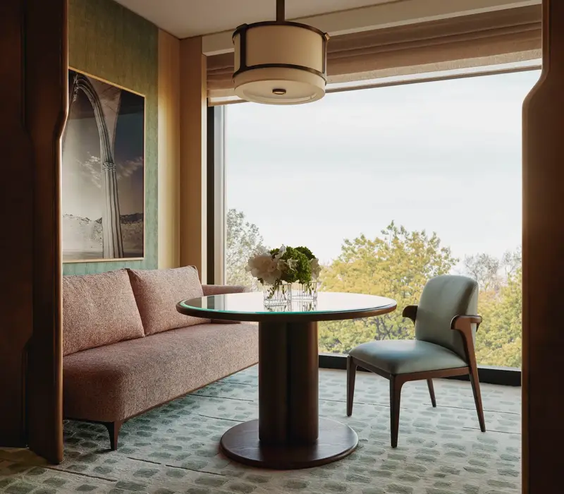 Modern dining nook with pink sofa, round glass table, and large window framing leafy treetop views 