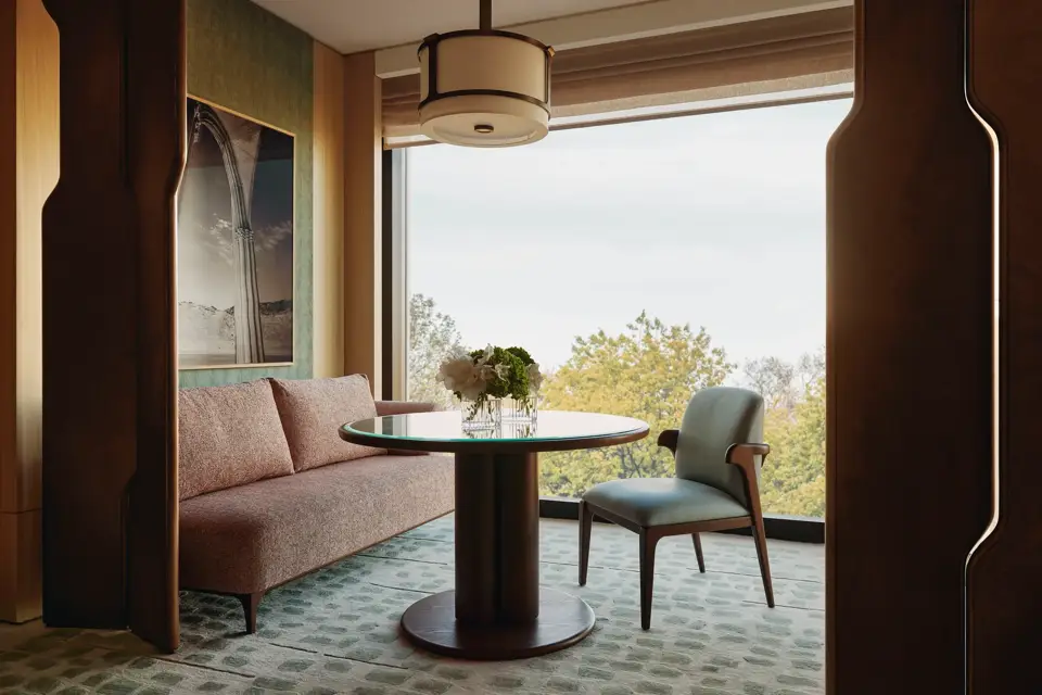 Modern dining nook with pink sofa, round glass table, and large window framing leafy treetop views 