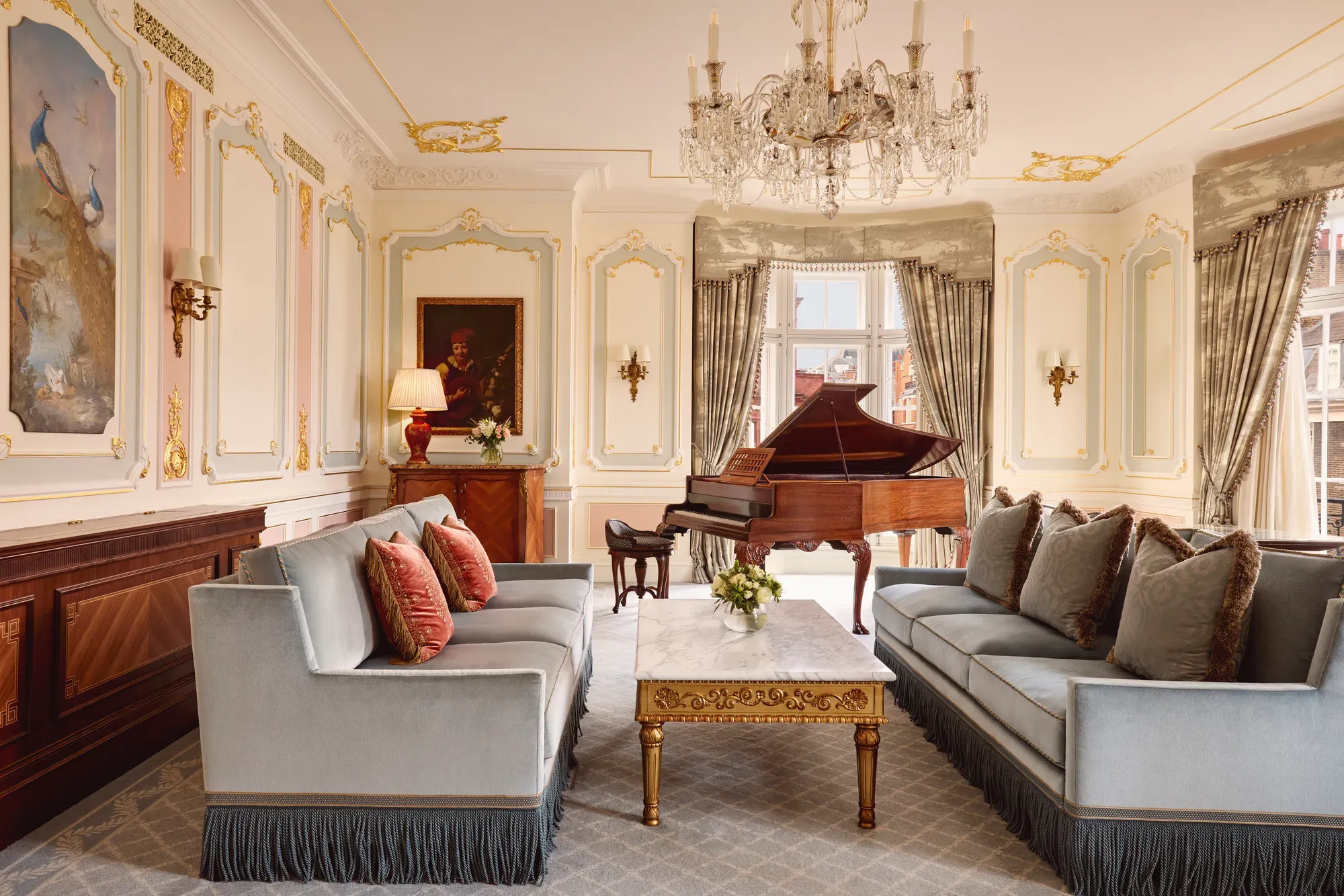 Opulent living room with two blue fringed sofas facing each other, a gold-detailed marble coffee table, and a grand piano beside tall curtained windows, illuminated by an ornate crystal chandelier.