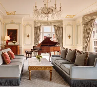 Opulent living room with two blue fringed sofas facing each other, a gold-detailed marble coffee table, and a grand piano beside tall curtained windows, illuminated by an ornate crystal chandelier.