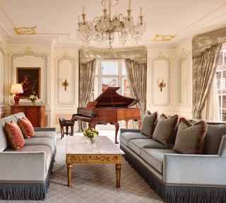 Opulent living room with two blue fringed sofas facing each other, a gold-detailed marble coffee table, and a grand piano beside tall curtained windows, illuminated by an ornate crystal chandelier.