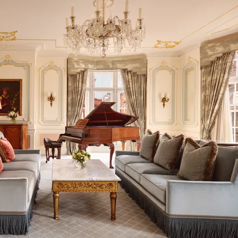 Opulent living room with two blue fringed sofas facing each other, a gold-detailed marble coffee table, and a grand piano beside tall curtained windows, illuminated by an ornate crystal chandelier.