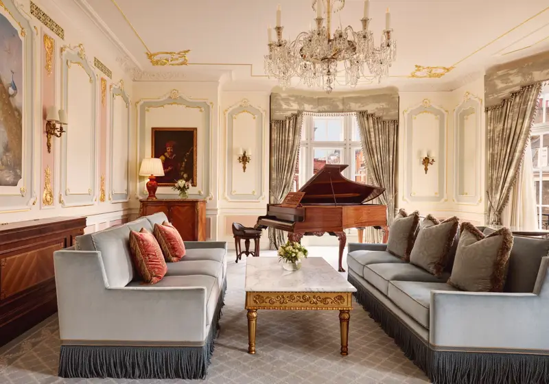 Opulent living room with two blue fringed sofas facing each other, a gold-detailed marble coffee table, and a grand piano beside tall curtained windows, illuminated by an ornate crystal chandelier.