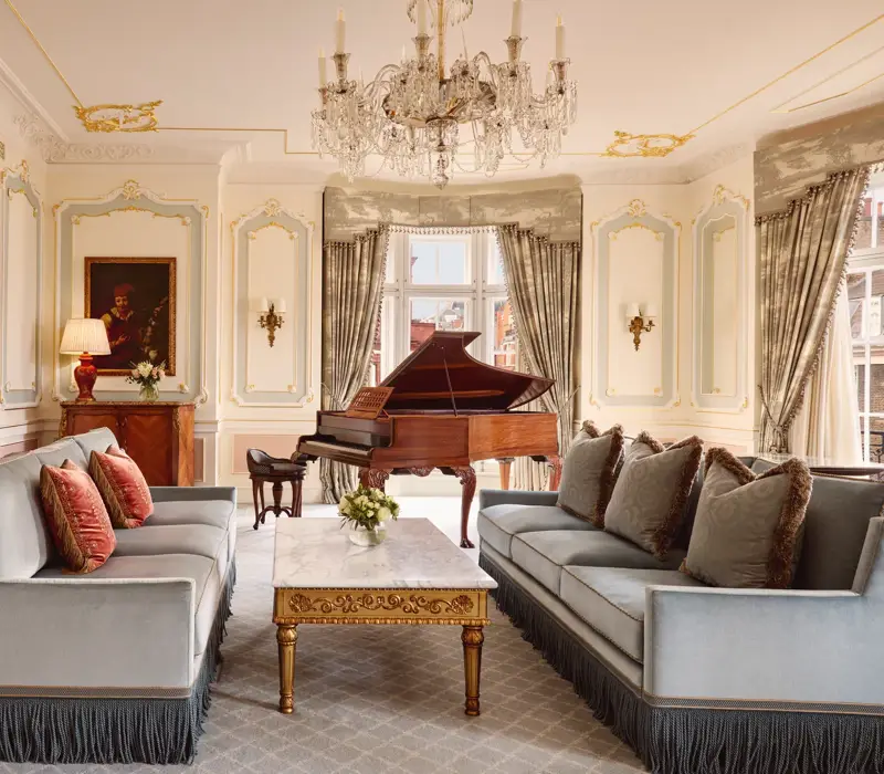 Opulent living room with two blue fringed sofas facing each other, a gold-detailed marble coffee table, and a grand piano beside tall curtained windows, illuminated by an ornate crystal chandelier.