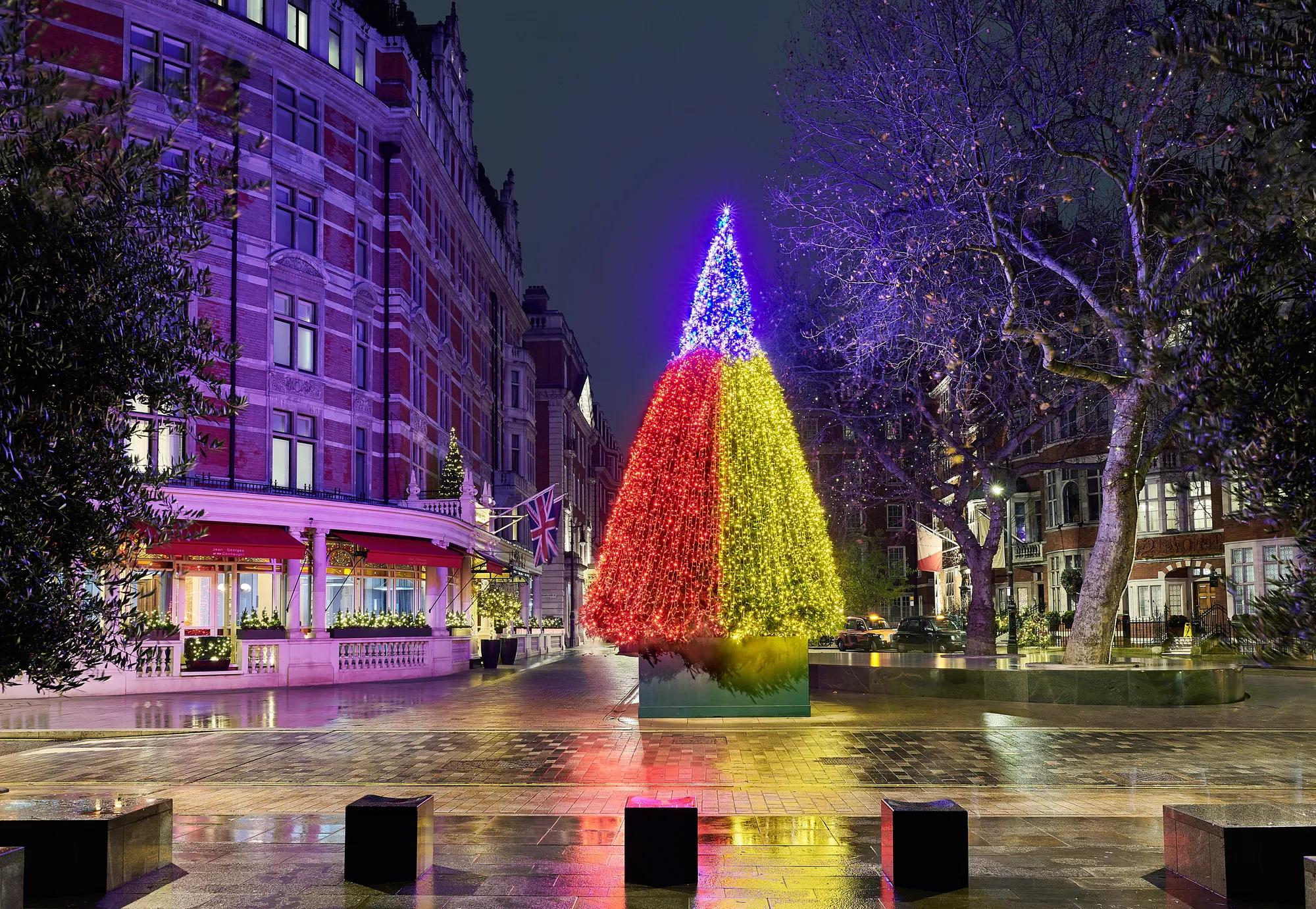 Multicoloured illuminated Christmas tree outside grand hotel on wet London street at night.