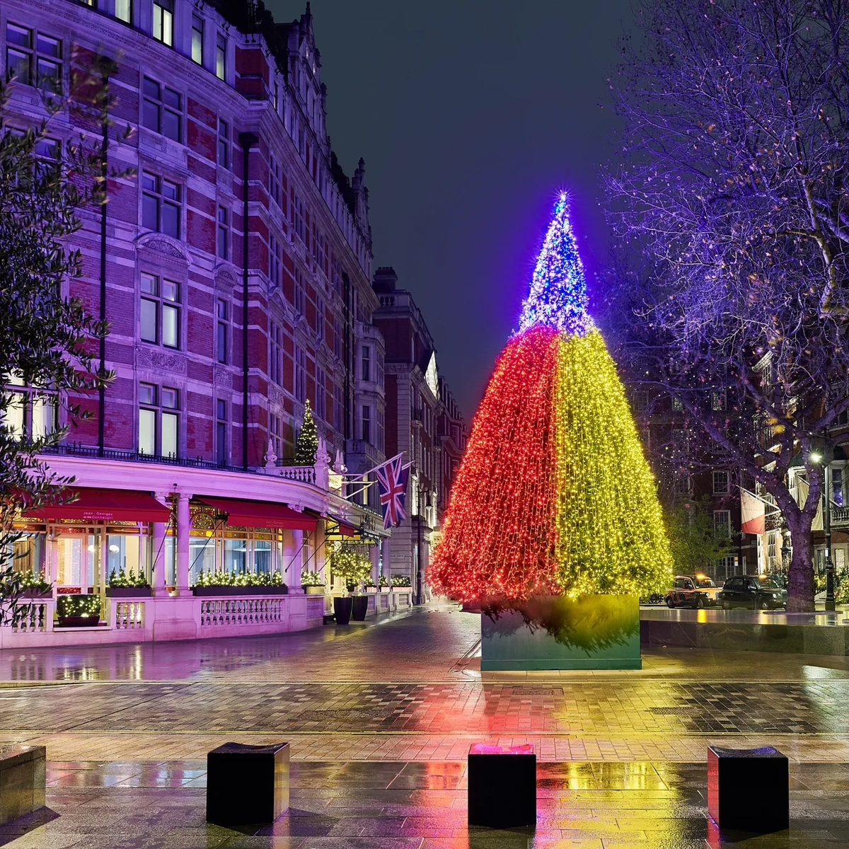 Multicoloured illuminated Christmas tree outside grand hotel on wet London street at night.