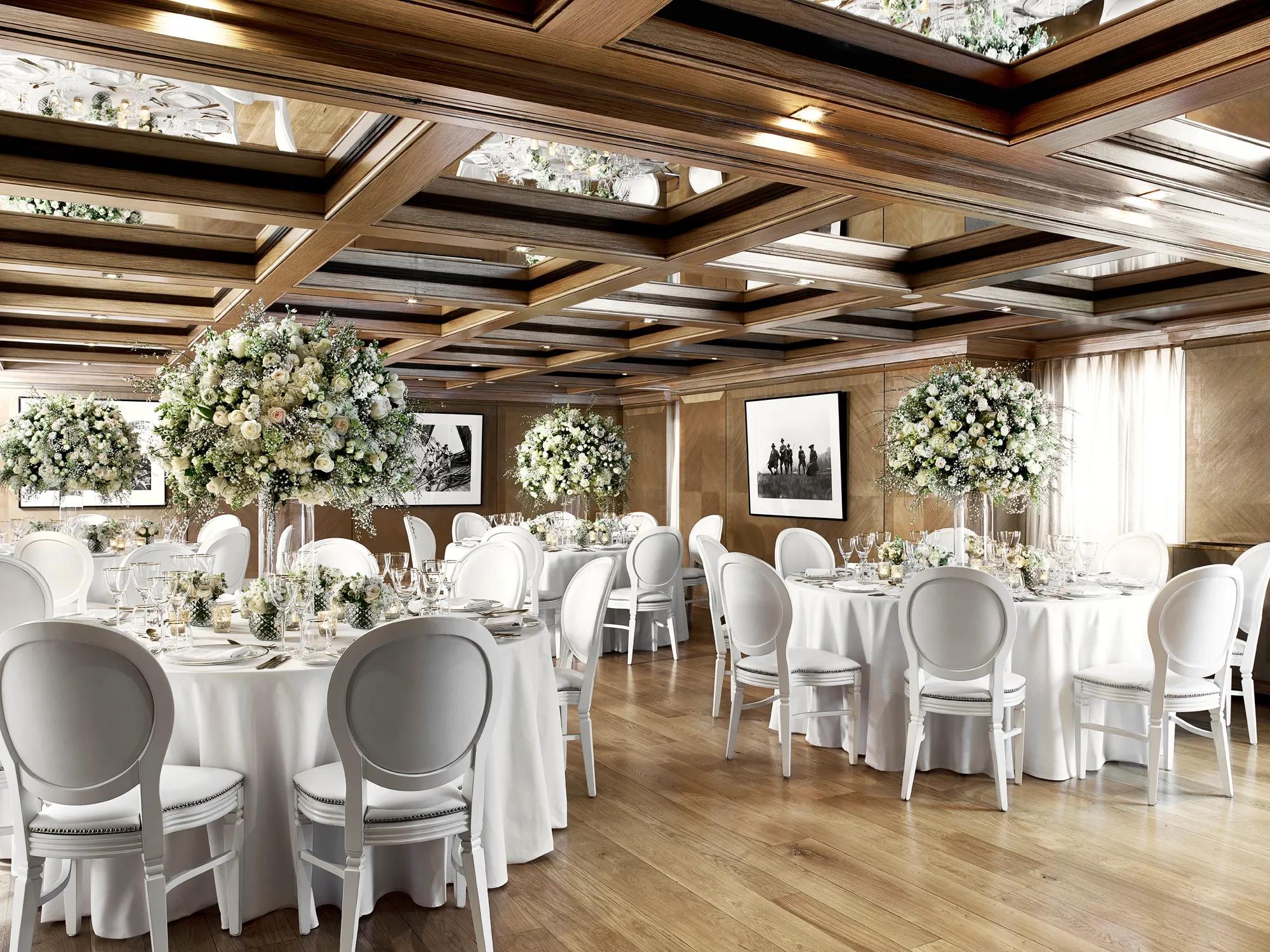 Elegant wedding dining room with round tables, white chairs, and tall floral centrepieces under a mirrored wooden ceiling.