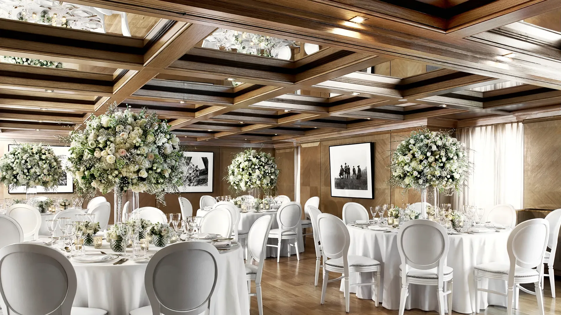 Elegant wedding dining room with round tables, white chairs, and tall floral centrepieces under a mirrored wooden ceiling.