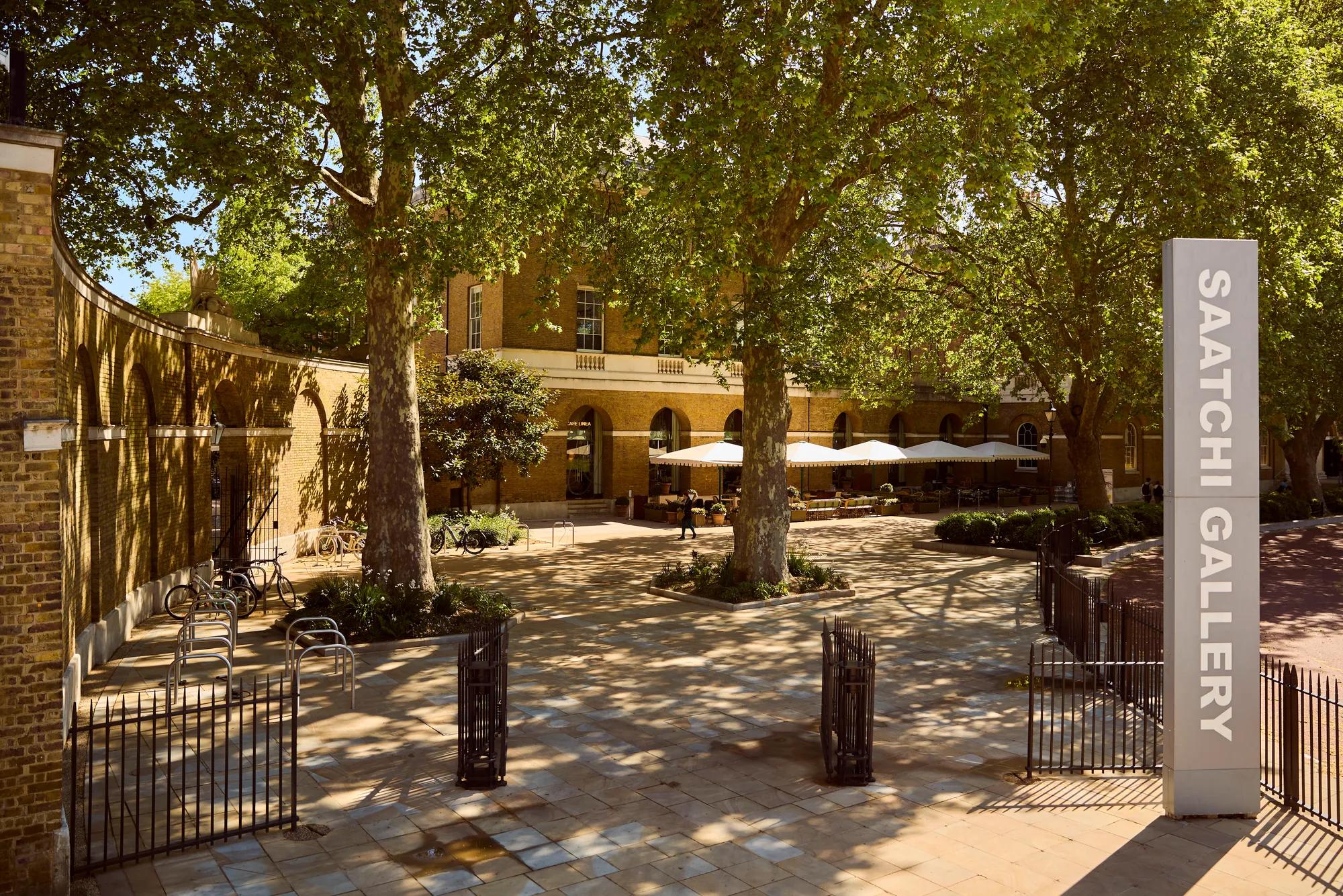 Entrance to Saatchi Gallery courtyard, shaded by trees, with outdoor seating and a vertical gallery sign.