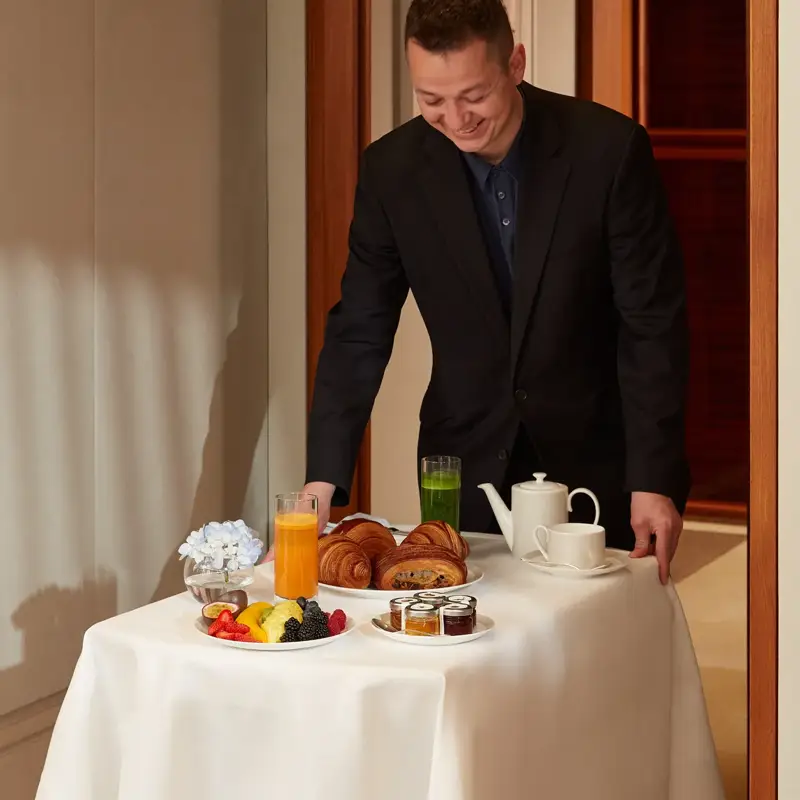 Hotel staff delivering breakfast with croissants, fruit, juices, and tea on a white-linen table in a suite hallway.