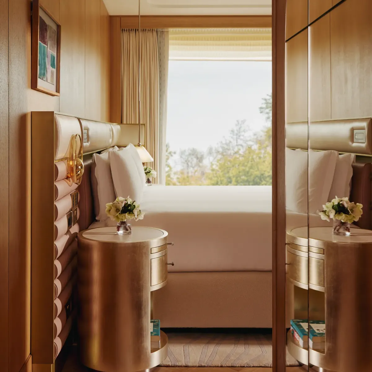 Detail of luxury suite bedroom with mirrored wardrobe, gold bedside tables, and window view over trees.