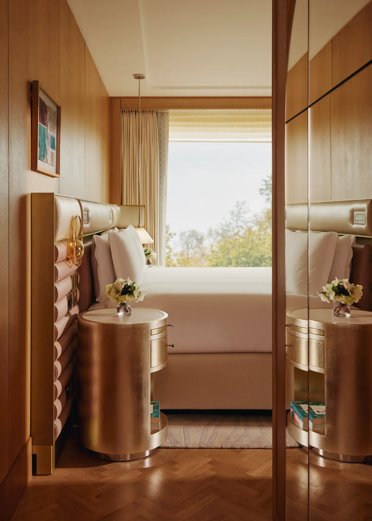 Detail of luxury suite bedroom with mirrored wardrobe, gold bedside tables, and window view over trees.