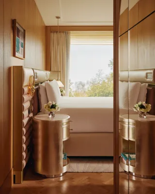 Detail of luxury suite bedroom with mirrored wardrobe, gold bedside tables, and window view over trees.