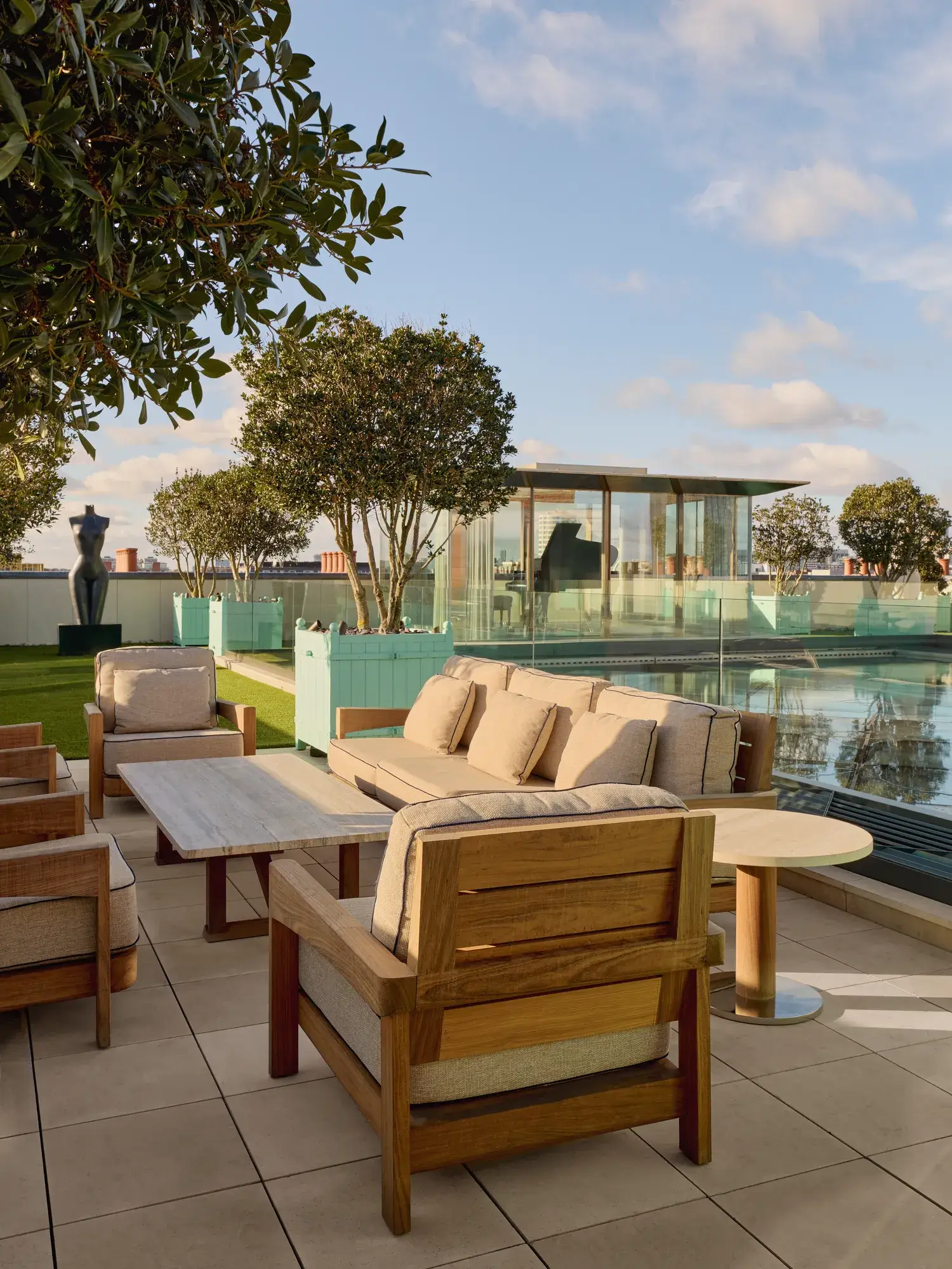 Rooftop terrace at Claridge’s with cushioned wooden lounge chairs, glass-edged pool, and a glass pavilion in the background.