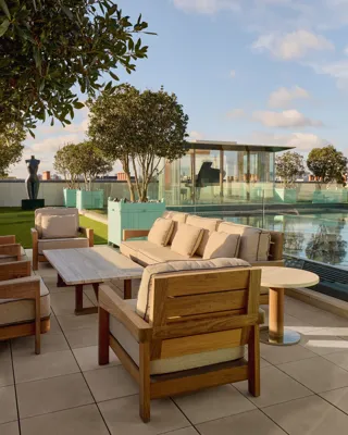 Rooftop terrace at Claridge’s with cushioned wooden lounge chairs, glass-edged pool, and a glass pavilion in the background.
