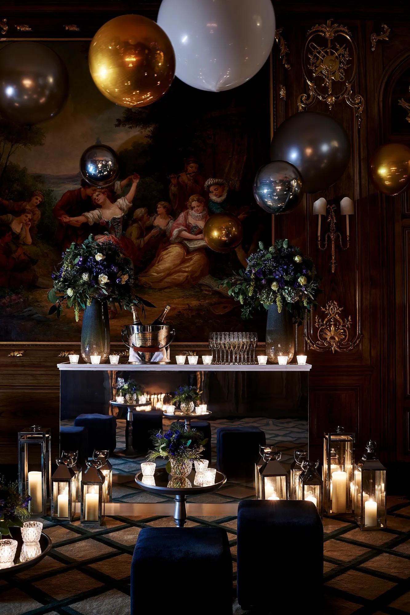 Elegant candlelit reception table with floral arrangements, lanterns, and metallic balloons set against ornate painted walls.