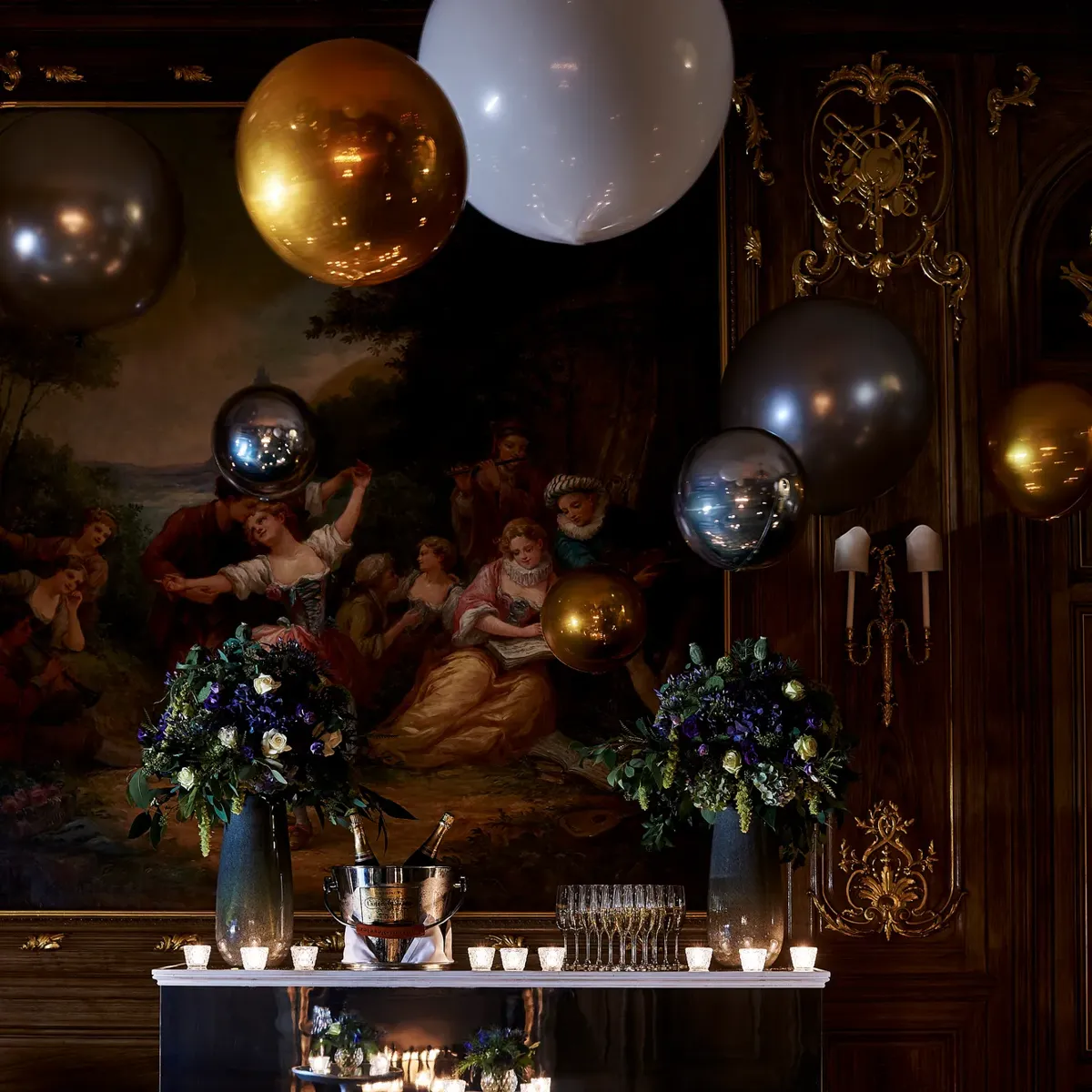 Elegant candlelit reception table with floral arrangements, lanterns, and metallic balloons set against ornate painted walls.