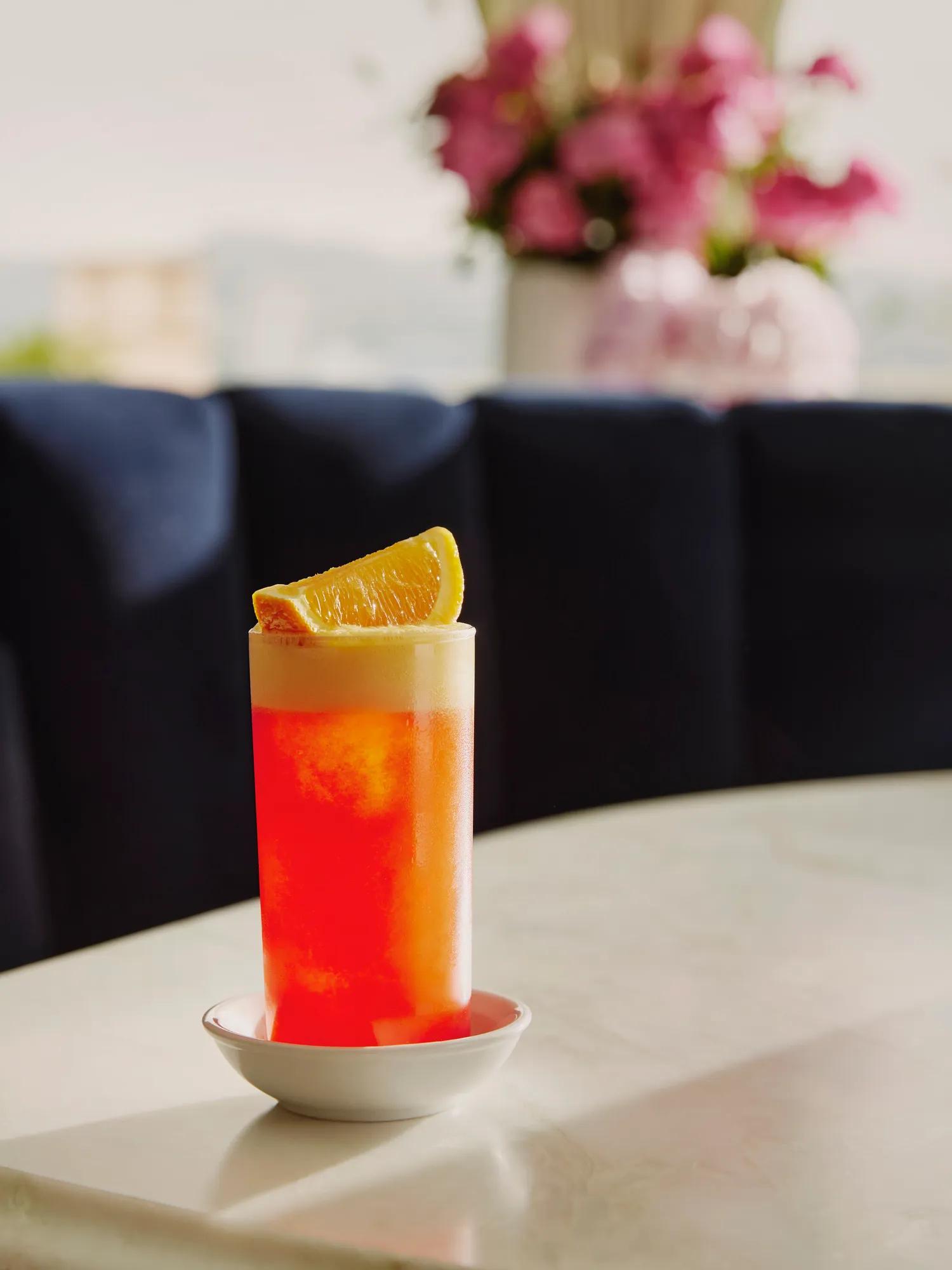 Tall orange-campari a highball with foamy head and an orange wedge on a marble table.