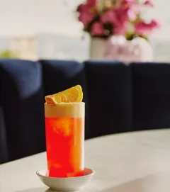 Tall orange-campari a highball with foamy head and an orange wedge on a marble table.