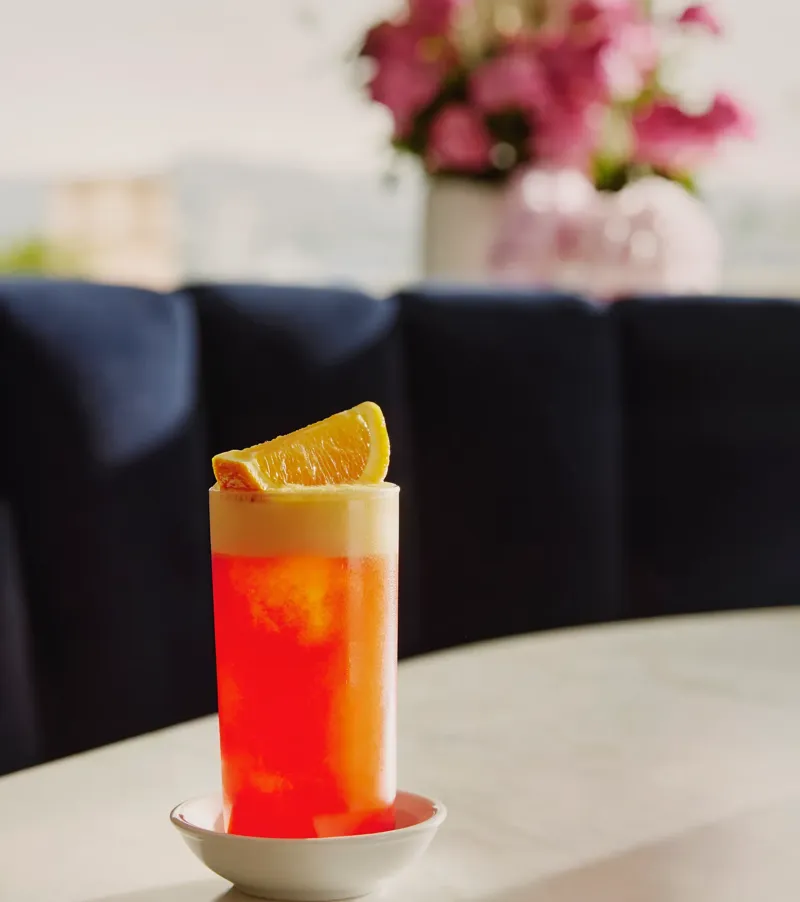 Tall orange-campari a highball with foamy head and an orange wedge on a marble table.
