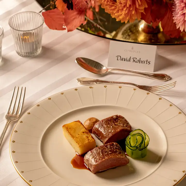 Plated fine dining dish at Claridge’s featuring seared meat, layered potato, and cucumber ribbons on an elegant cream and gold-rimmed plate set on a white tablecloth.