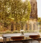 An outdoor terrace dining area with a stone table and two modern chairs, overlooking a historic church tower and surrounded by leafy trees.