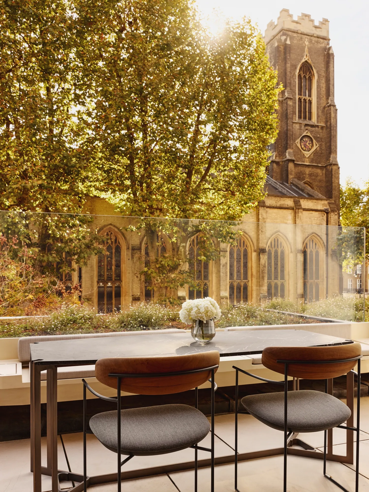 An outdoor terrace dining area with a stone table and two modern chairs, overlooking a historic church tower and surrounded by leafy trees.