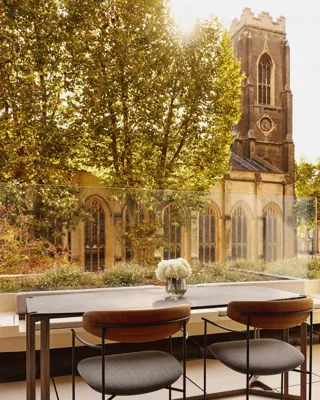An outdoor terrace dining area with a stone table and two modern chairs, overlooking a historic church tower and surrounded by leafy trees.