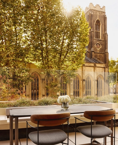 An outdoor terrace dining area with a stone table and two modern chairs, overlooking a historic church tower and surrounded by leafy trees.