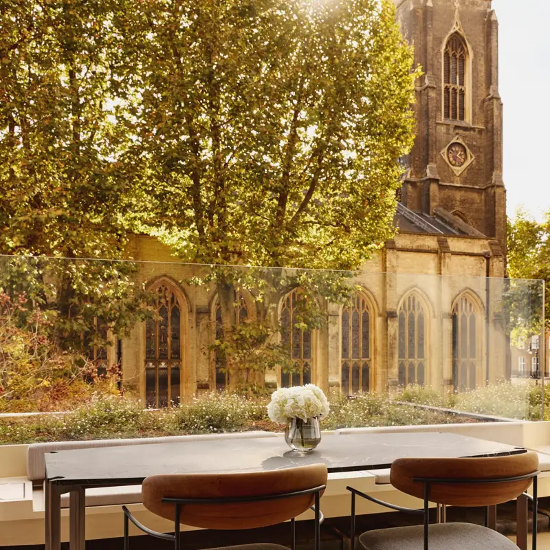 An outdoor terrace dining area with a stone table and two modern chairs, overlooking a historic church tower and surrounded by leafy trees.