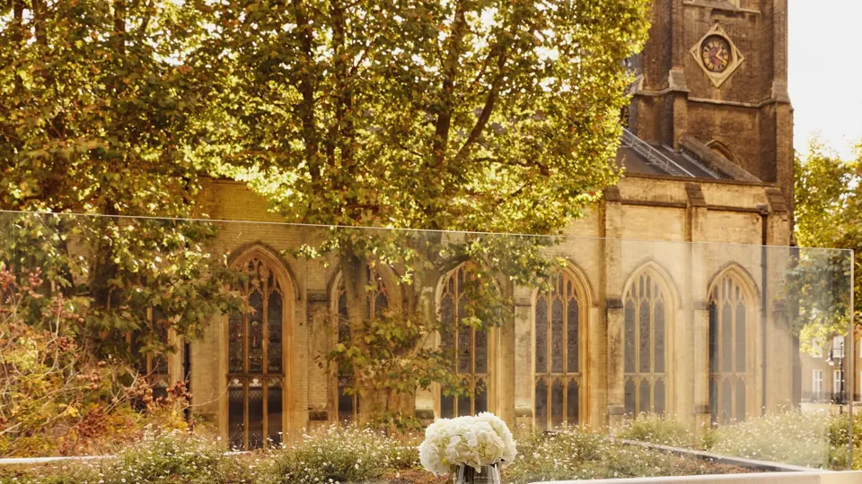 An outdoor terrace dining area with a stone table and two modern chairs, overlooking a historic church tower and surrounded by leafy trees.