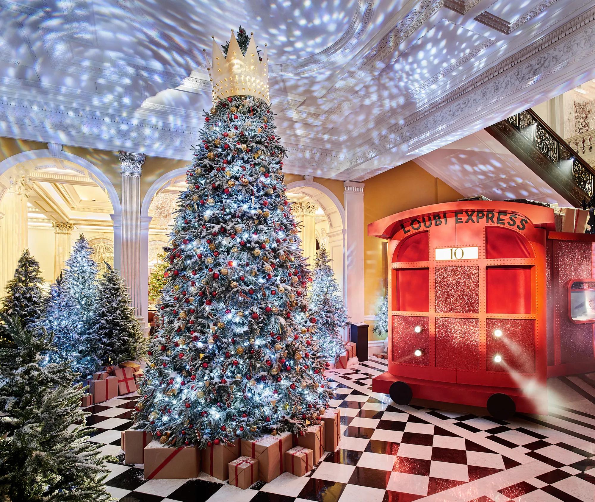 Grand festive lobby with large decorated Christmas tree topped by crown, surrounded by gifts, beside a red train installation on checkered marble floor.