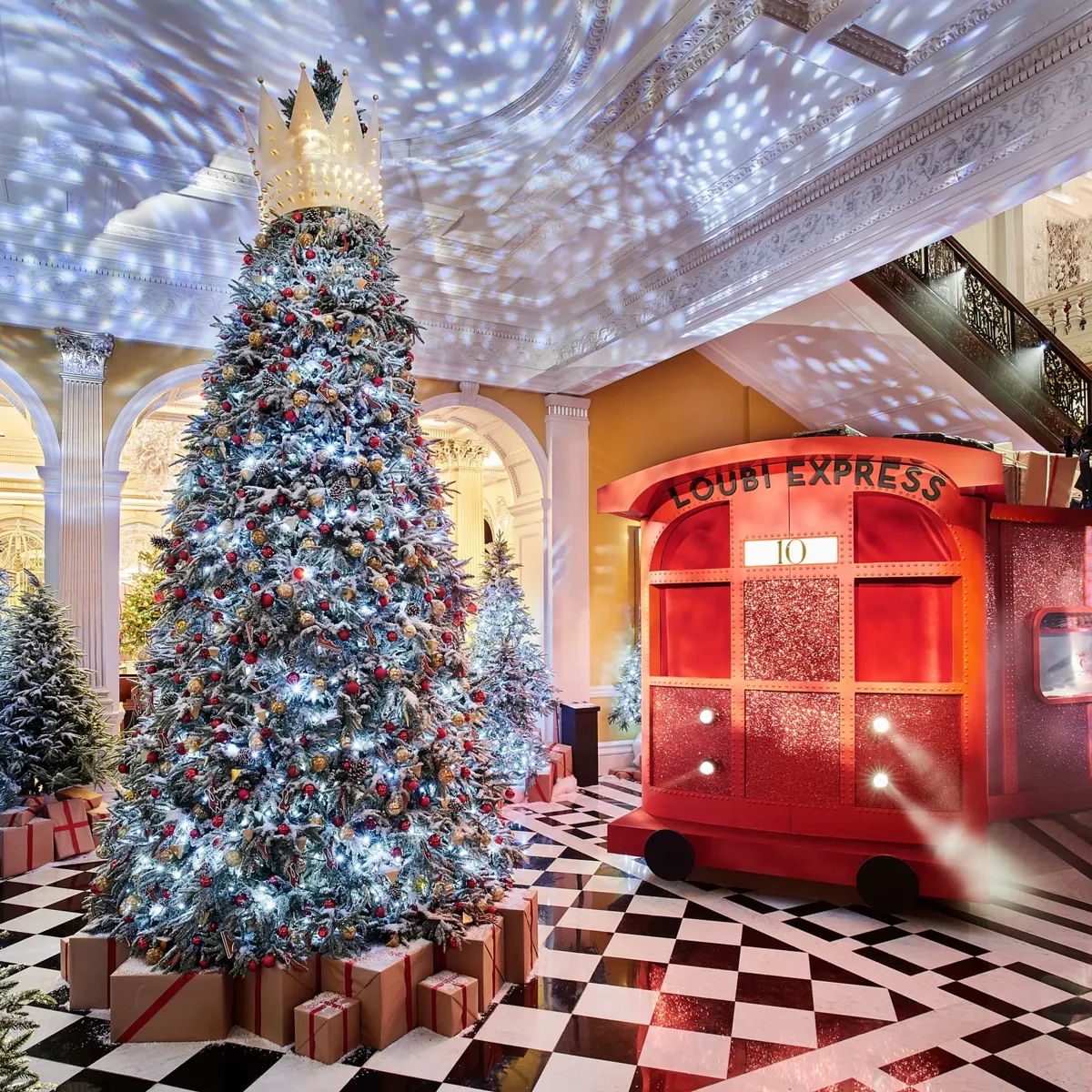 Grand festive lobby with large decorated Christmas tree topped by crown, surrounded by gifts, beside a red train installation on checkered marble floor.