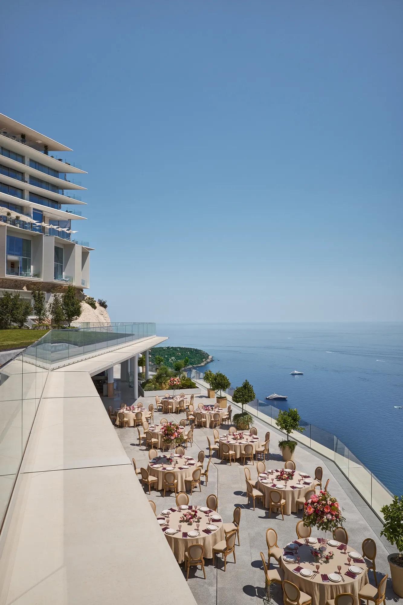 Installation événementielle en terrasse avec des tables rondes décorées de grands centres de table floraux, surplombant la mer avec des yachts au loin, à côté d’un hôtel moderne.