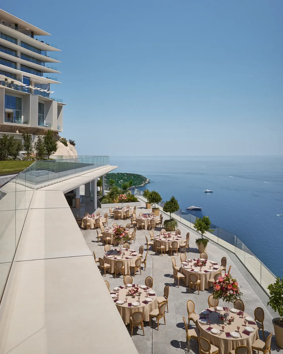 Installation événementielle en terrasse avec des tables rondes décorées de grands centres de table floraux, surplombant la mer avec des yachts au loin, à côté d’un hôtel moderne.