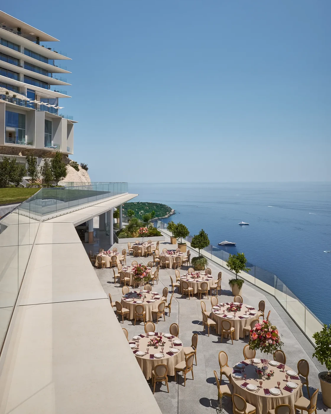 Installation événementielle en terrasse avec des tables rondes décorées de grands centres de table floraux, surplombant la mer avec des yachts au loin, à côté d’un hôtel moderne.