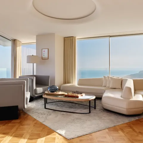 Modern living room with curved white sofa, grey armchairs, and coffee table, featuring large windows with sea and mountain views.
