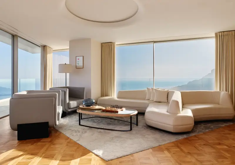 Modern living room with curved white sofa, grey armchairs, and coffee table, featuring large windows with sea and mountain views.
