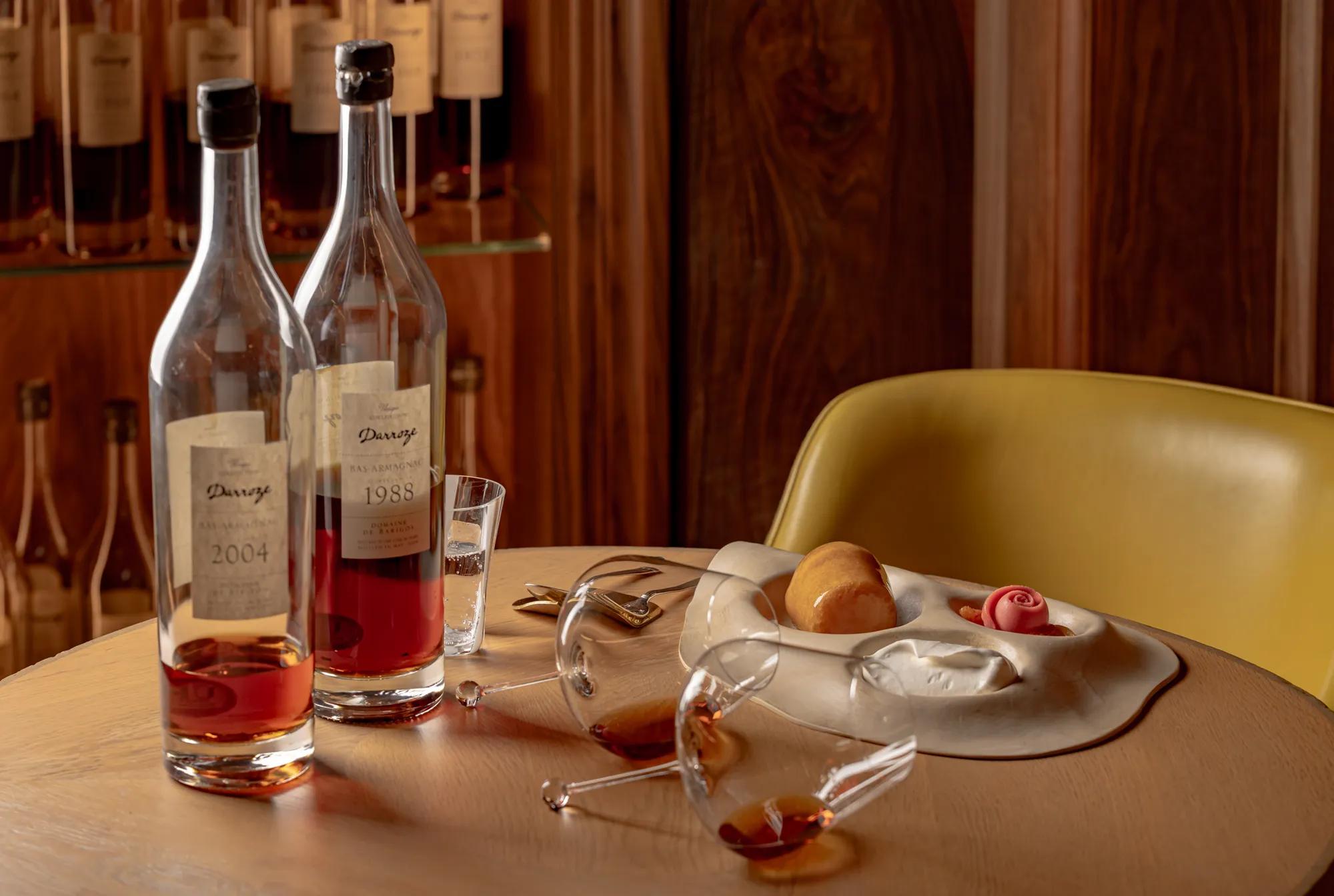 Two bottles of amber spirits with glasses on a wooden table, alongside plated dessert and a yellow chair in a warm interior