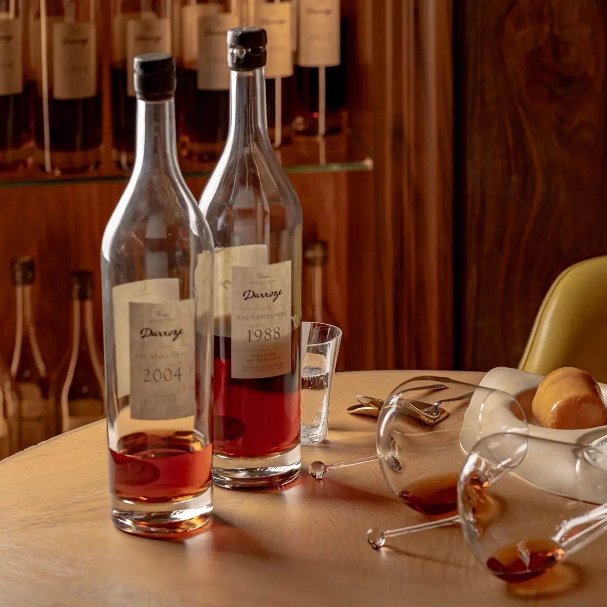 Two bottles of amber spirits with glasses on a wooden table, alongside plated dessert and a yellow chair in a warm interior