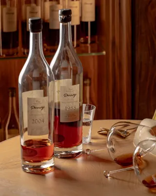 Two bottles of amber spirits with glasses on a wooden table, alongside plated dessert and a yellow chair in a warm interior