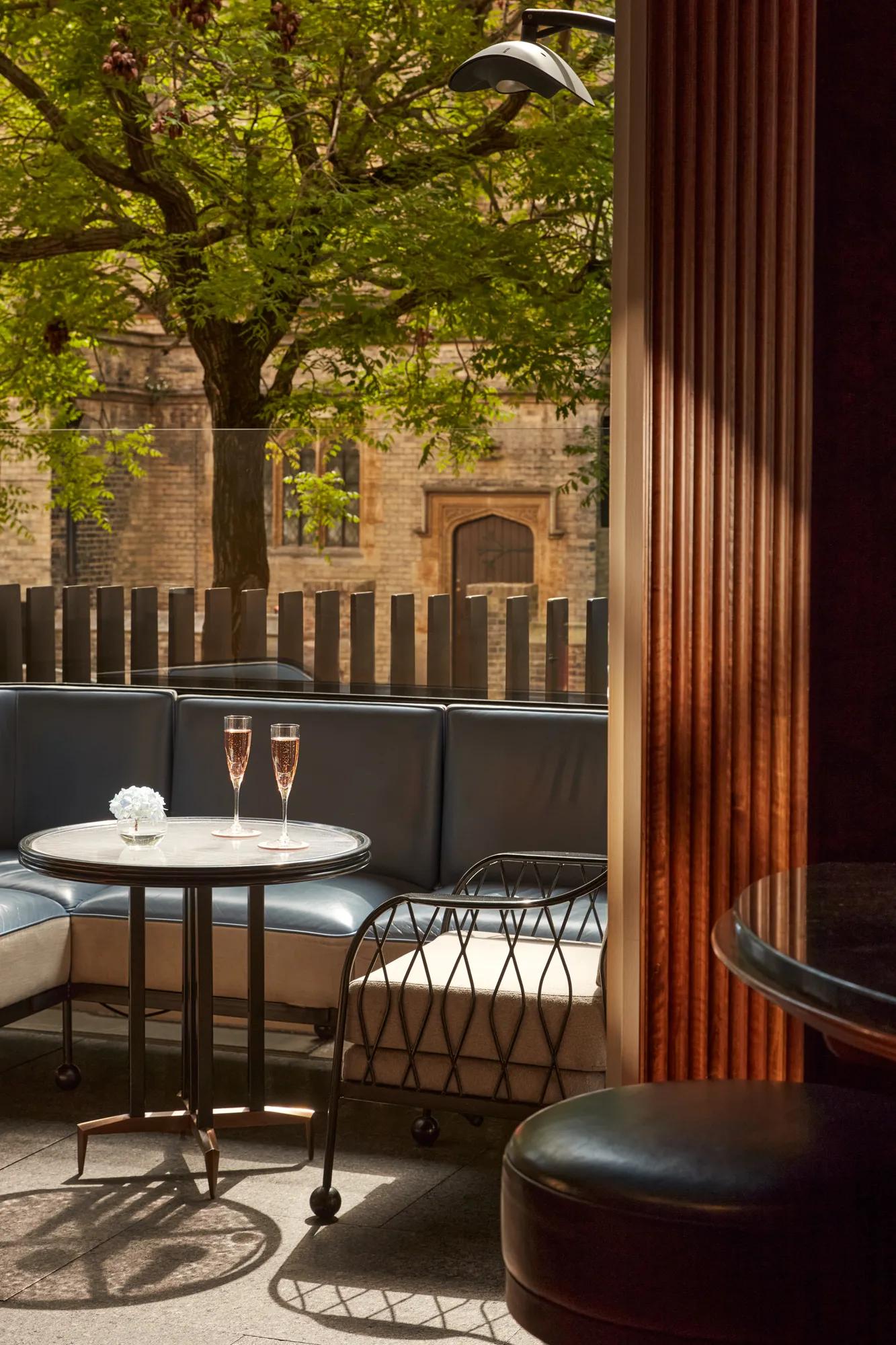 Outdoor terrace seating with two champagne flutes on a round table, shaded by trees and overlooking historic stone architecture