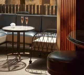 Outdoor terrace seating with two champagne flutes on a round table, shaded by trees and overlooking historic stone architecture