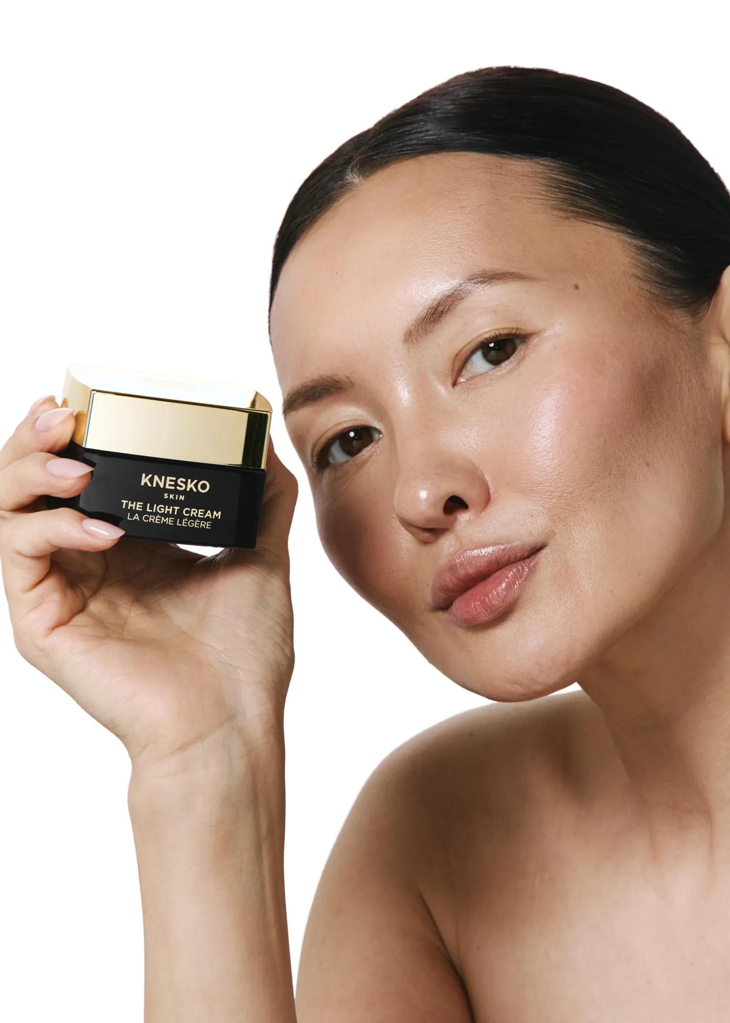 Woman holding a jar of KNESKO Skin The Light Cream near face against a plain light background