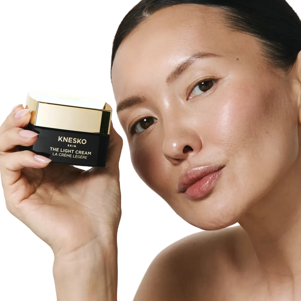 Woman holding a jar of KNESKO Skin The Light Cream near face against a plain light background