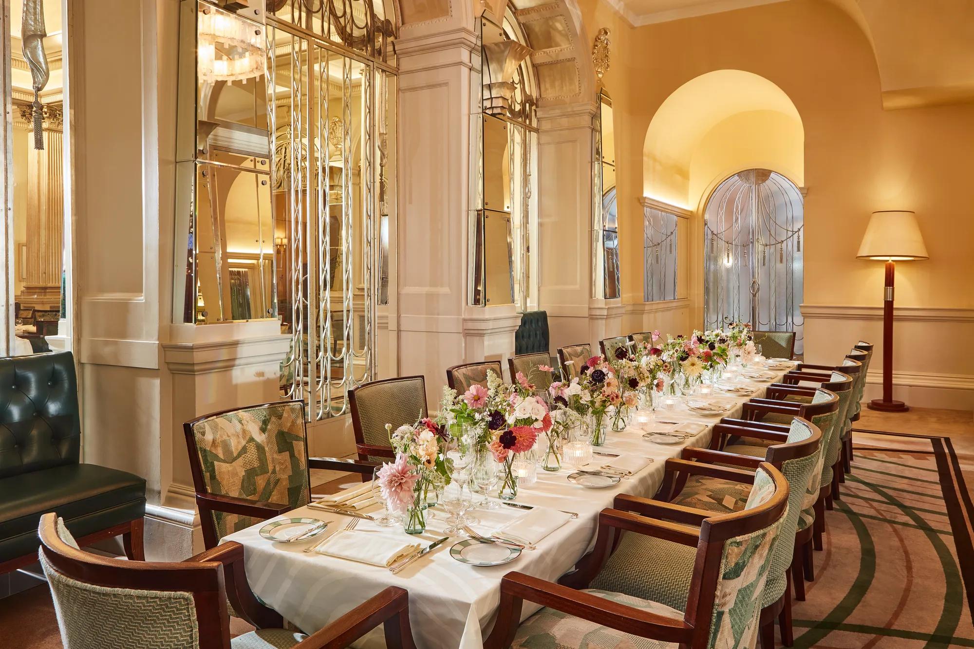 Elegant private dining room at Claridge’s featuring a long table set with white linens, floral centrepieces, and vintage-style chairs beneath mirrored Art Deco arches.