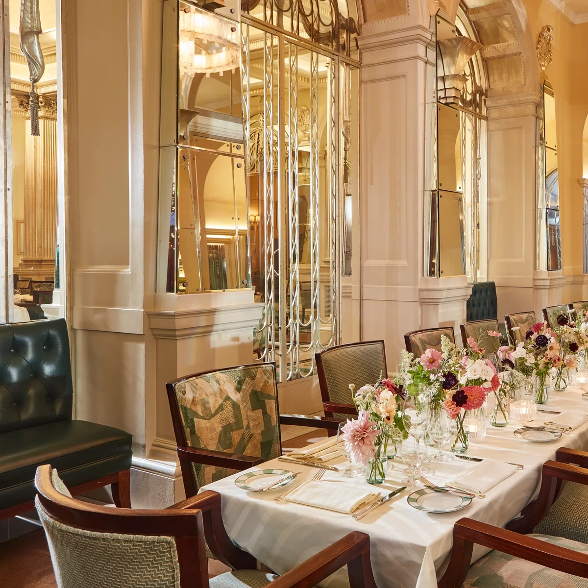Elegant private dining room at Claridge’s featuring a long table set with white linens, floral centrepieces, and vintage-style chairs beneath mirrored Art Deco arches.